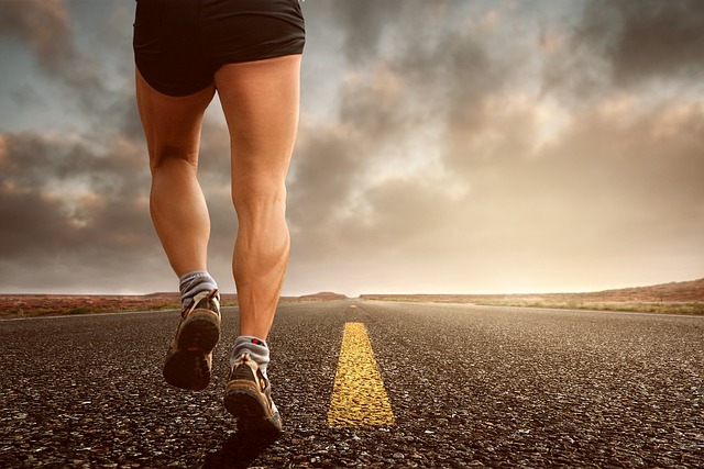 A person wearing athletic shorts and running shoes is running down an empty asphalt road with a yellow center line, heading towards a horizon under a cloudy sky. The perspective is from behind, focusing on their legs and the open road ahead.