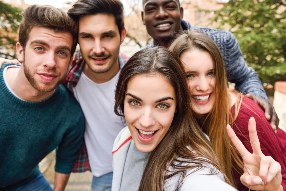 A group of five friends smiling and posing for a selfie outdoors. The person in front is making a peace sign with their fingers. Trees and the blurred background of a street can be seen behind them. Everyone appears happy and close together.