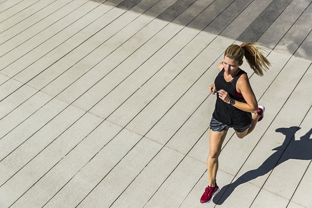 A woman with a ponytail is jogging on a wide, paved surface. She is wearing a black tank top, gray shorts, and red running shoes. The photo is taken from an overhead angle, capturing her shadow on the ground.