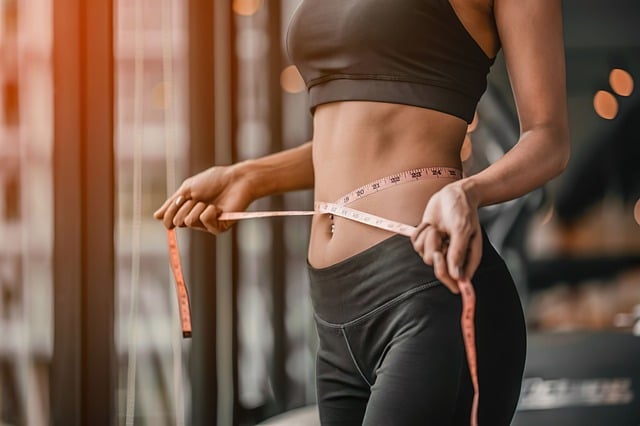 A fit individual wearing a black sports bra and leggings measures their waist with a tape measure. They stand in a bright room with large windows, suggesting a gym or fitness studio. The focus is on the person's toned midsection and the tape measure.